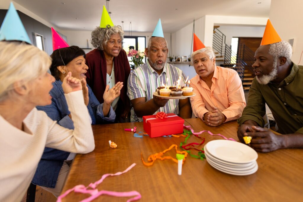 Multiracial senior friends wearing party hats looking at man blowing candles in nursing home