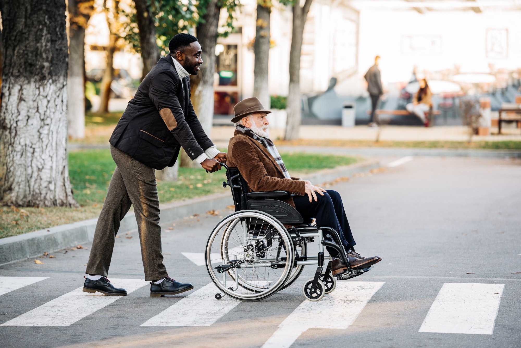 side view of senior disabled man in wheelchair and young african american cuidador riding by street
